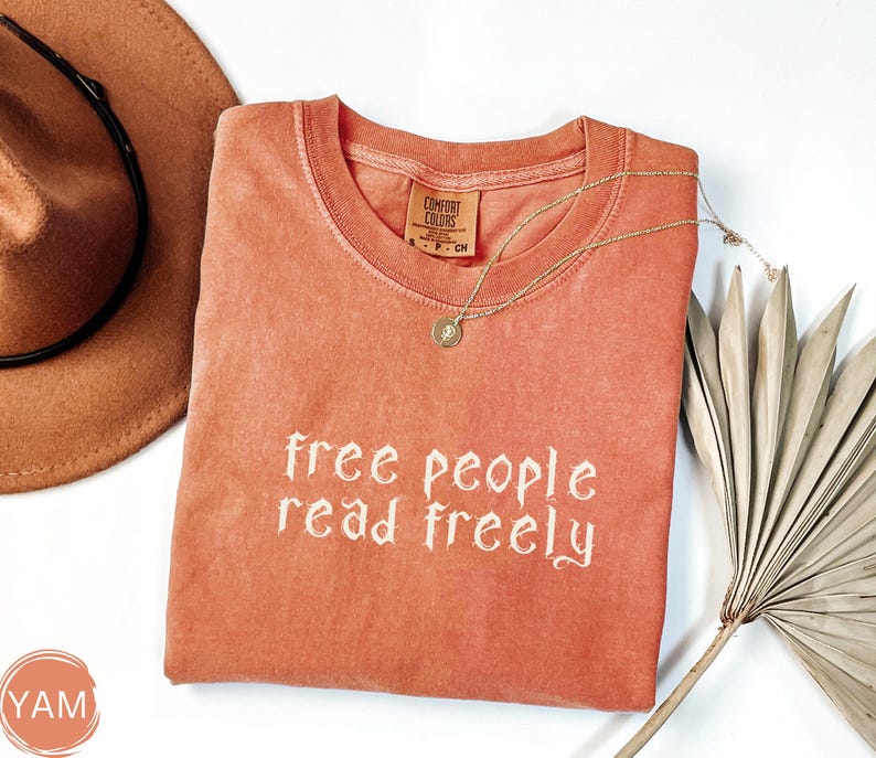 May include: A coral-colored t-shirt with the phrase "free people read freely" in white script. A gold necklace with a pendant rests on the shirt. A brown hat and dried palm leaves are also in the image.