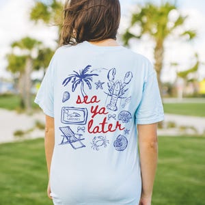 Puede incluir: Una camiseta azul claro con un dise&ntilde;o gr&aacute;fico de una palmera, una langosta, una concha, un cangrejo, una estrella de mar, una lata de sardinas, una silla de playa y el texto "sea ya later".