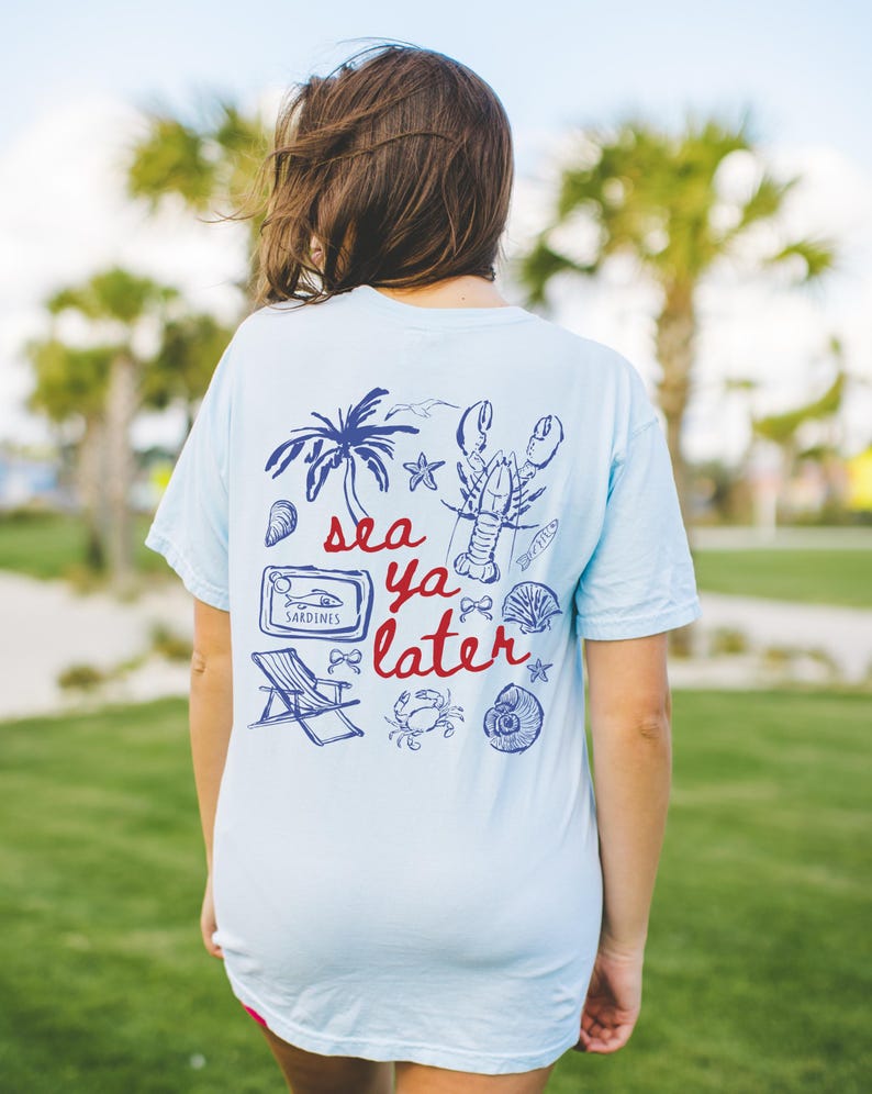 Puede incluir: Una camiseta azul claro con un dise&ntilde;o gr&aacute;fico de una palmera, una langosta, una concha, un cangrejo, una estrella de mar, una lata de sardinas, una silla de playa y el texto "sea ya later".