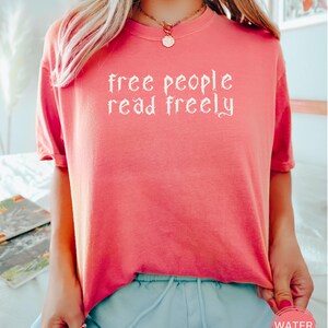 May include: Coral-colored t-shirt with the text "free people read freely" in a gothic font. The shirt has short sleeves and a round neckline. A small, round sticker with the word "WATERMELON" is visible on the bottom right.