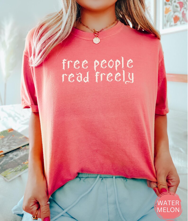 May include: Coral-colored t-shirt with the text "free people read freely" in a gothic font. The shirt has short sleeves and a round neckline. A small, round sticker with the word "WATERMELON" is visible on the bottom right.