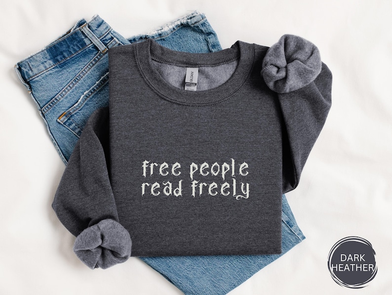 May include: Dark heather gray sweatshirt with the words 'free people read freely' in white, stylized text. The sweatshirt is folded over a pair of blue jeans. The sleeves are cuffed. A dark heather circle with the words 'DARK HEATHER' is in the bottom right corner.