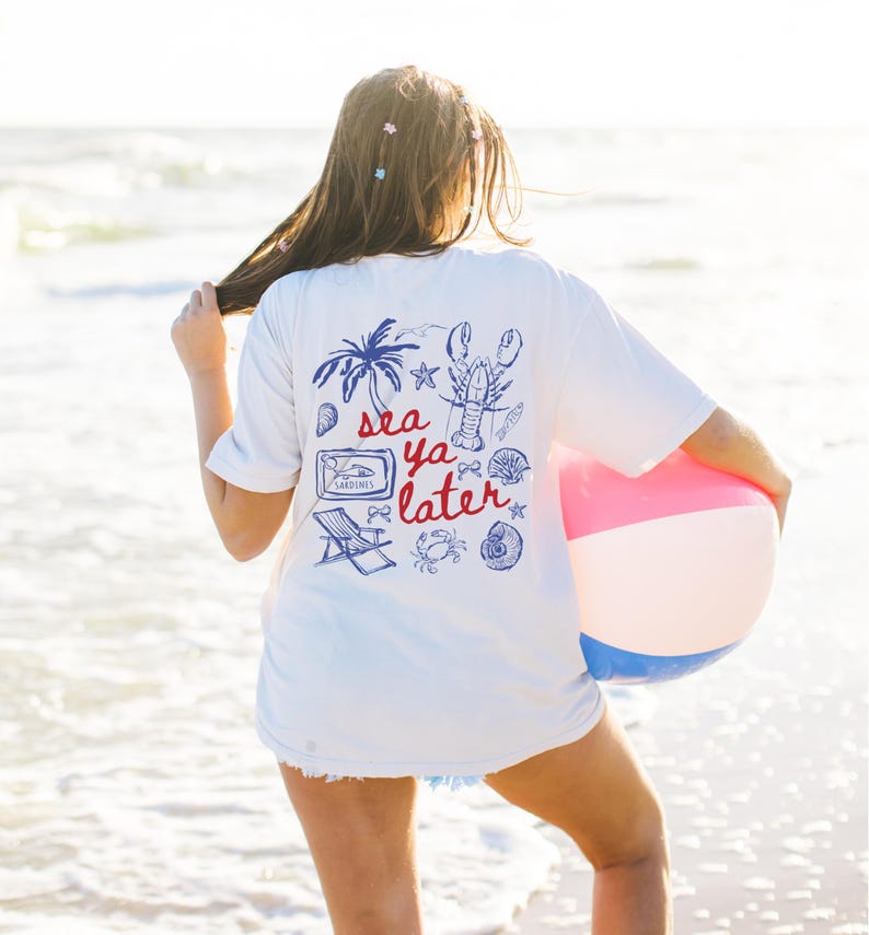 May include: A white t-shirt with a blue and red graphic print that says 'sea ya later'. The graphic includes a lobster, a palm tree, a beach chair, a seashell, a crab, and a can of sardines.
