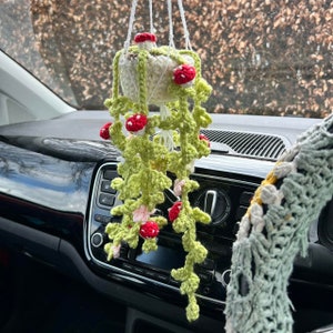 Op de afbeelding: Een gehaakte hangplant met een witte pot en groene bladeren. De plant heeft rode en roze bloemen en hangt aan de achteruitkijkspiegel van een auto.