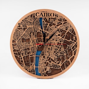 May include: A wooden clock with a laser-cut design of the city of Cairo, Egypt. The clock face features the Nile River in blue, with the city streets and landmarks in brown. The words "CAIRO" and "GIZA" are visible on the clock face.