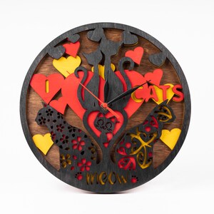 May include: A wooden clock with a black background and red, yellow, and black cat silhouettes. The clock face has black hands and the words "I Love Cats" and "Meow" are cut out of the wood.