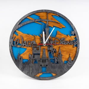 Puede incluir: Un reloj de pared de madera con una silueta del Castillo de Cenicienta con el texto "Walt Disney World" en una fuente estilizada. La esfera del reloj es azul con un fondo gris y agujas blancas.