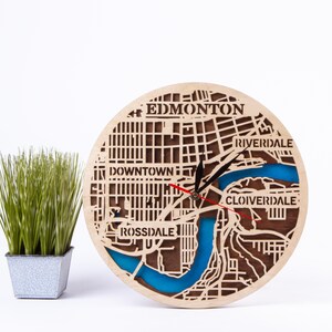 May include: A wooden clock featuring a map of Edmonton, Alberta, Canada. The clock face is made of wood and has a blue river running through the city. The clock hands are black and red. The clock is 30 cm in diameter.