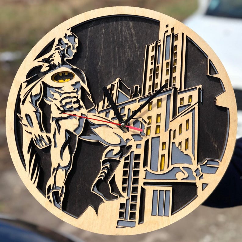 Pu&ograve; includere: Un orologio da parete in legno con una silhouette di Batman che calcia un edificio. Il quadrante dell'orologio &egrave; nero con lancette bianche e un logo Batman giallo.