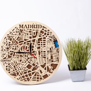 May include: A wooden clock with a map of Madrid, Spain, on the face. The clock hands are black and red, and the clock face is surrounded by a light brown wooden frame. The words "MADRID" and "CENTRO" are visible on the clock face.