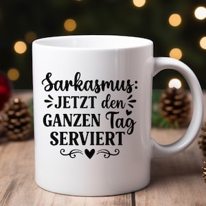 May include: White ceramic mug with the German text "Sarkasmus: JETZT den GANZEN Tag SERVIERT" in black script. The mug features a handle and decorative flourishes. The mug is likely used for hot beverages.