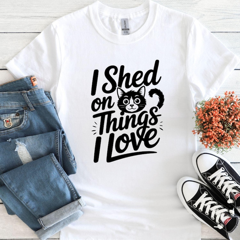 May include: White t-shirt with the text "I Shed on Things I Love" and a black cat graphic. The shirt is paired with blue jeans and black and white sneakers. A small bouquet of orange flowers is also present.