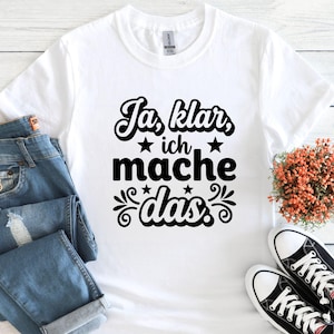 May include: White t-shirt with the German phrase "Ja, klar, ich mache das" in black script. The design includes stars and decorative flourishes. The shirt is paired with blue jeans and black and white sneakers.