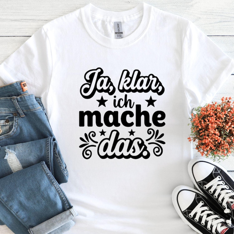 May include: White t-shirt with the German phrase "Ja, klar, ich mache das" in black script. The design includes stars and decorative flourishes. The shirt is paired with blue jeans and black and white sneakers.