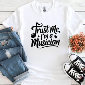 May include: White t-shirt with the text "Trust Me, I'm a Musician" in a black, cursive font. The shirt is paired with blue jeans and black and white sneakers. A small bouquet of orange flowers is also present.