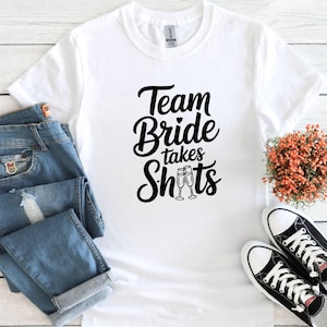 May include: White t-shirt with black text that reads "Team Bride takes Shots" with a heart and two champagne glasses.
