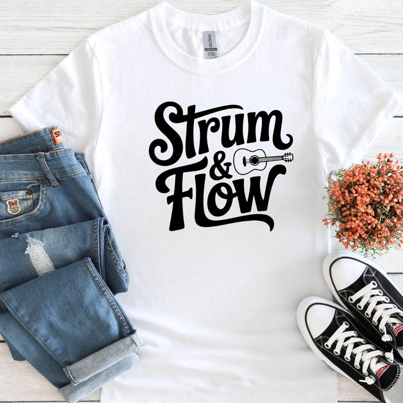 May include: White t-shirt with the text "Strum & Flow" in a black, stylized font, accompanied by a guitar graphic. The shirt is paired with blue jeans and black and white sneakers, suggesting a casual, musical theme.