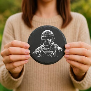 May include: A round, black slate coaster featuring a detailed white illustration of a soldier in uniform, complete with a helmet and tactical gear. The coaster is held up by a person.