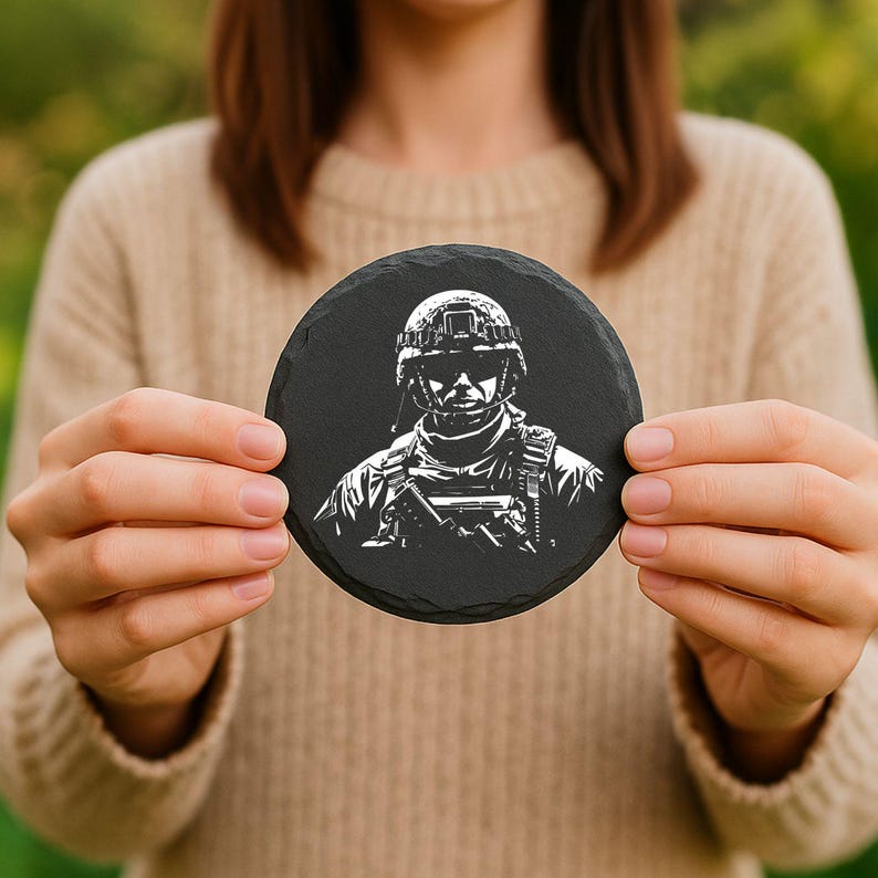 May include: A round, black slate coaster featuring a detailed white illustration of a soldier in uniform, complete with a helmet and tactical gear. The coaster is held up by a person.