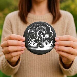 May include: A round, black slate coaster featuring a detailed white illustration of a dragon facing a robed figure. The dragon has large wings and is positioned above the robed figure. The coaster is held by a person.