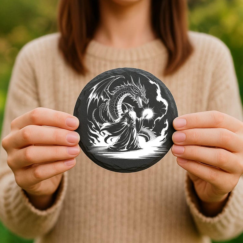 May include: A round, black slate coaster featuring a detailed white illustration of a dragon facing a robed figure. The dragon has large wings and is positioned above the robed figure. The coaster is held by a person.