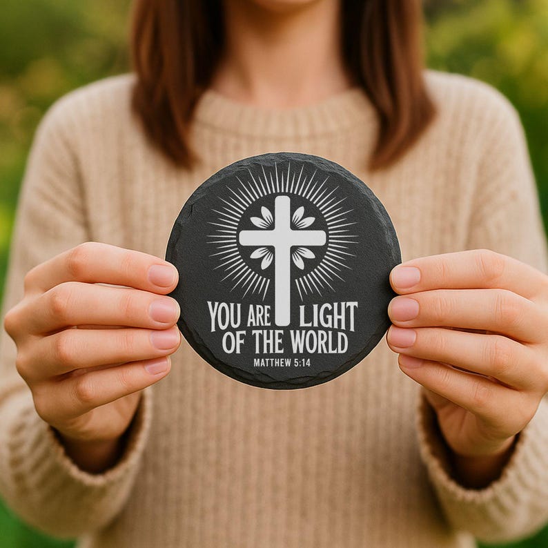 Puede incluir: Un posavasos redondo de pizarra negra con un dise&ntilde;o de cruz blanca y el texto "YOU ARE LIGHT OF THE WORLD" y "MATTHEW 5:14". El posavasos est&aacute; sostenido por una persona que lleva un jersey beige.