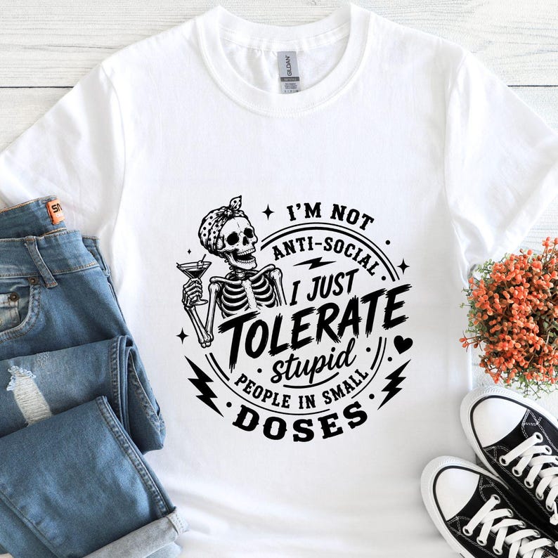 K&ouml;nnte beinhalten: Wei&szlig;es T-Shirt mit einem schwarzen Grafikdesign eines Skeletts mit Cocktail und dem Text: "I'M NOT ANTI-SOCIAL I JUST TOLERATE stupid PEOPLE IN SMALL DOSES." Das Shirt wird mit blauen Jeans und schwarzen Turnschuhen kombiniert.