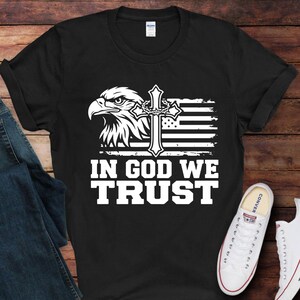 May include: Black t-shirt featuring a white graphic of an eagle's head, a cross, and a stylized American flag. The text "IN GOD WE TRUST" is printed in bold white letters below the graphic. A pair of white sneakers is visible in the bottom right corner.