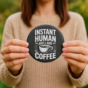 May include: A round, black slate coaster with the text "INSTANT HUMAN JUST ADD COFFEE" in white. A white coffee cup graphic is centered below the text. The coaster is held by a person.