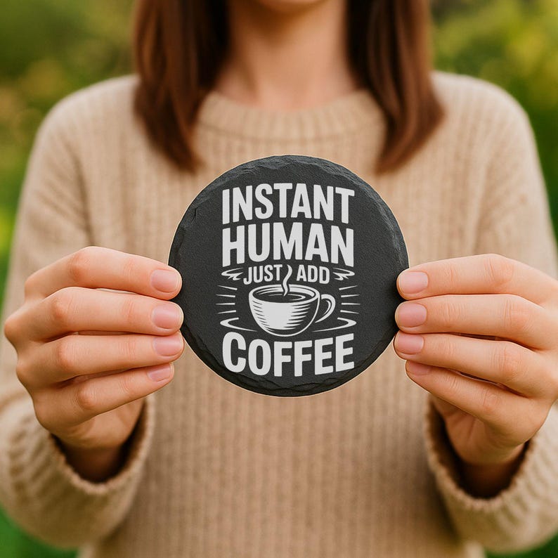 May include: A round, black slate coaster with the text "INSTANT HUMAN JUST ADD COFFEE" in white. A white coffee cup graphic is centered below the text. The coaster is held by a person.
