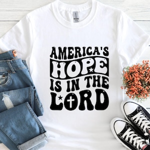 May include: White t-shirt with the text "AMERICA'S HOPE IS IN THE LORD" in a black, retro-style font. The shirt is paired with blue jeans and black and white sneakers.