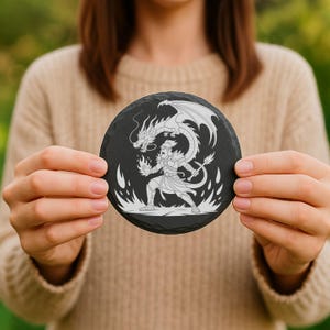 May include: A round, black slate coaster featuring a white illustration of a dragon and a warrior. The dragon is above the warrior, with flames around the warrior. The coaster is held by a person.