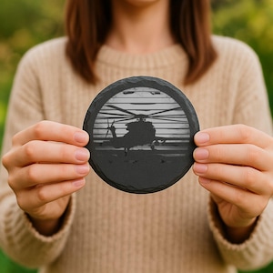 May include: A round, black slate coaster featuring a detailed silhouette of a helicopter against a striped background. The coaster is held up, showcasing its design and texture. The coaster is a decorative item.