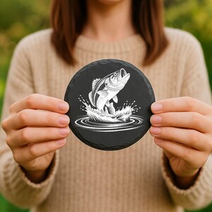 May include: A round, black slate coaster featuring a white graphic of a leaping fish with water splashes. The coaster is held up by a person. The background is blurred.
