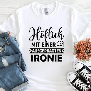 May include: White t-shirt with black text that reads "H&ouml;flich MIT EINER AUSGEPR&Auml;GTEN IRONIE" with a mustache and monocle graphic. The shirt is paired with blue jeans and black and white sneakers.