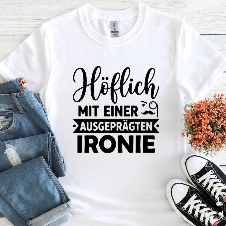 May include: White t-shirt with black text that reads "H&ouml;flich MIT EINER AUSGEPR&Auml;GTEN IRONIE" with a mustache and monocle graphic. The shirt is paired with blue jeans and black and white sneakers.