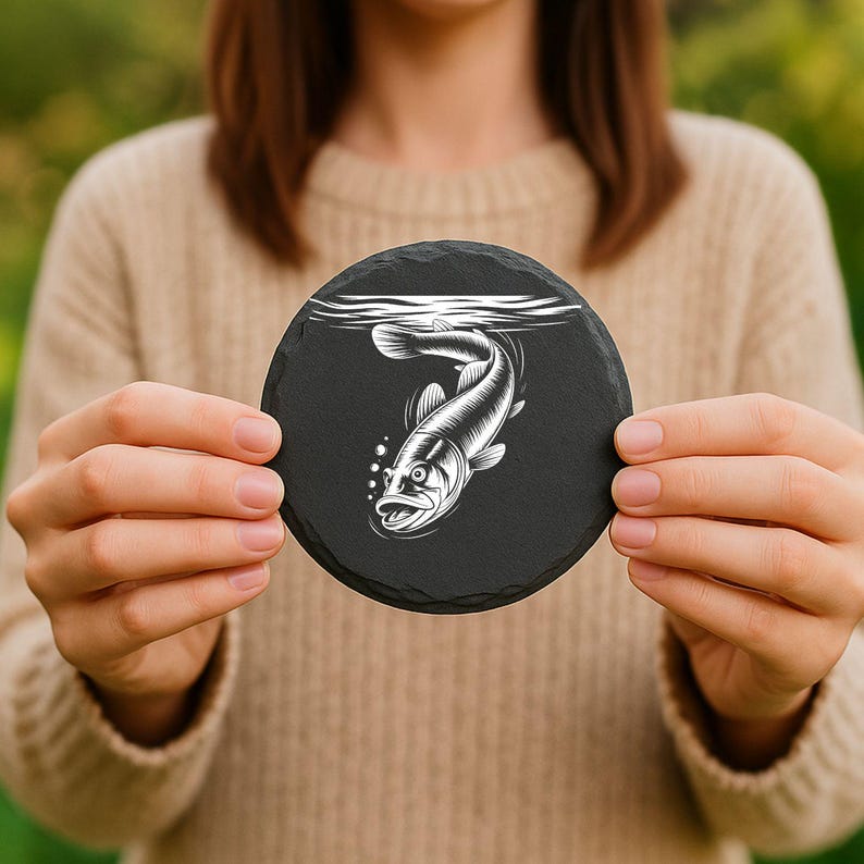 May include: A round, black slate coaster featuring a detailed white illustration of a fish swimming underwater. The fish is depicted with open mouth and bubbles. The coaster is held up by a person.