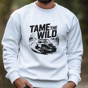 Puede incluir: Sudadera blanca con un dise&ntilde;o gr&aacute;fico negro de un veh&iacute;culo todoterreno y el texto "TAME THE WILD". El dise&ntilde;o muestra un veh&iacute;culo en un camino de tierra con monta&ntilde;as. La sudadera es de cuello redondo.