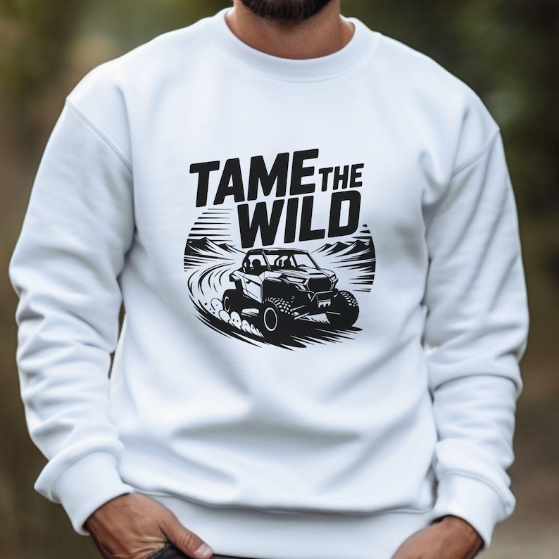 Puede incluir: Sudadera blanca con un dise&ntilde;o gr&aacute;fico negro de un veh&iacute;culo todoterreno y el texto "TAME THE WILD". El dise&ntilde;o muestra un veh&iacute;culo en un camino de tierra con monta&ntilde;as. La sudadera es de cuello redondo.