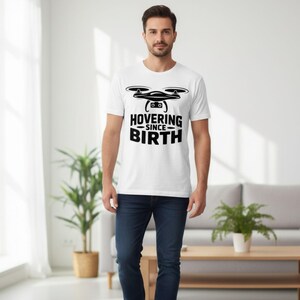 May include: White t-shirt with a black drone graphic and the text "HOVERING SINCE BIRTH". The shirt is made of a soft material and is suitable for casual wear. The person in the image is wearing blue jeans.