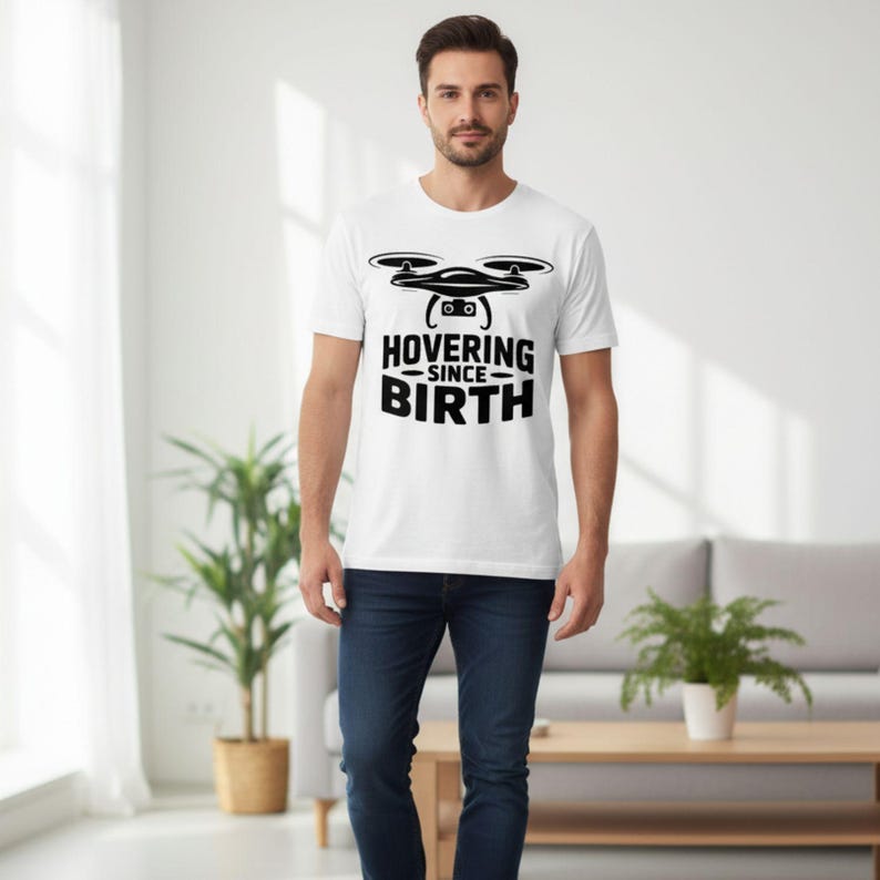 May include: White t-shirt with a black drone graphic and the text "HOVERING SINCE BIRTH". The shirt is made of a soft material and is suitable for casual wear. The person in the image is wearing blue jeans.