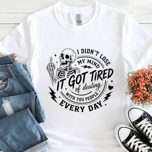 K&ouml;nnte beinhalten: Wei&szlig;es T-Shirt mit einem schwarzen Skelett-Motiv, das den Mittelfinger zeigt, mit dem Text "I didn't lose my mind, it got tired of dealing with you people every day." Das Shirt wird mit blauen Jeans und schwarzen Turnschuhen gezeigt.
