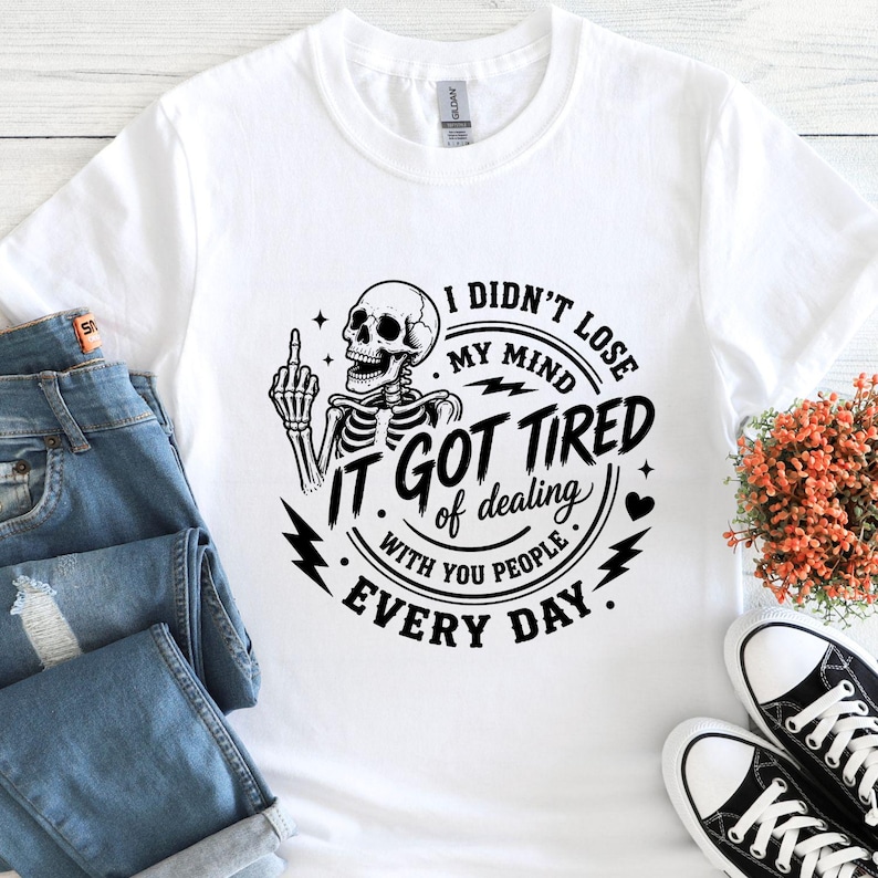 K&ouml;nnte beinhalten: Wei&szlig;es T-Shirt mit einem schwarzen Skelett-Motiv, das den Mittelfinger zeigt, mit dem Text "I didn't lose my mind, it got tired of dealing with you people every day." Das Shirt wird mit blauen Jeans und schwarzen Turnschuhen gezeigt.