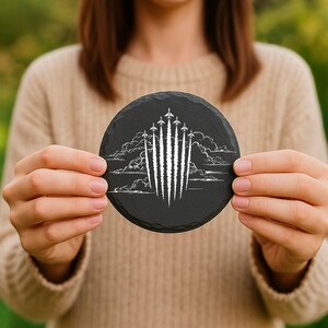 May include: A round, black slate coaster featuring a white etched design of several jet airplanes ascending through clouds. The coaster is held up by a person, showcasing the detailed artwork and the natural texture of the slate.