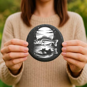 May include: A round, black slate coaster featuring a detailed white illustration of a fish jumping out of the water, with a reflection below. The design includes a full moon and stars in the background. The coaster is held up by a person.