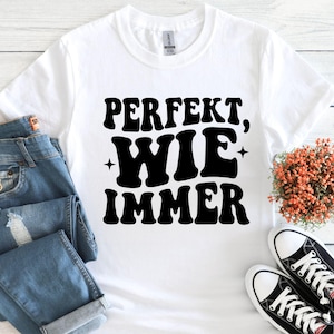 May include: White t-shirt with the text "PERFEKT, WIE IMMER" in bold black letters. The shirt is paired with blue jeans and black and white sneakers. A small bouquet of orange flowers is also visible.