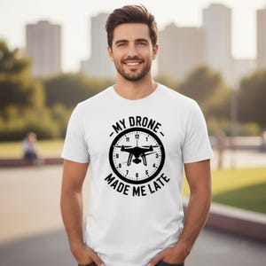 May include: White t-shirt with a black graphic of a drone inside a clock face. The text around the clock reads "MY DRONE MADE ME LATE". The shirt is short-sleeved and worn by a person.