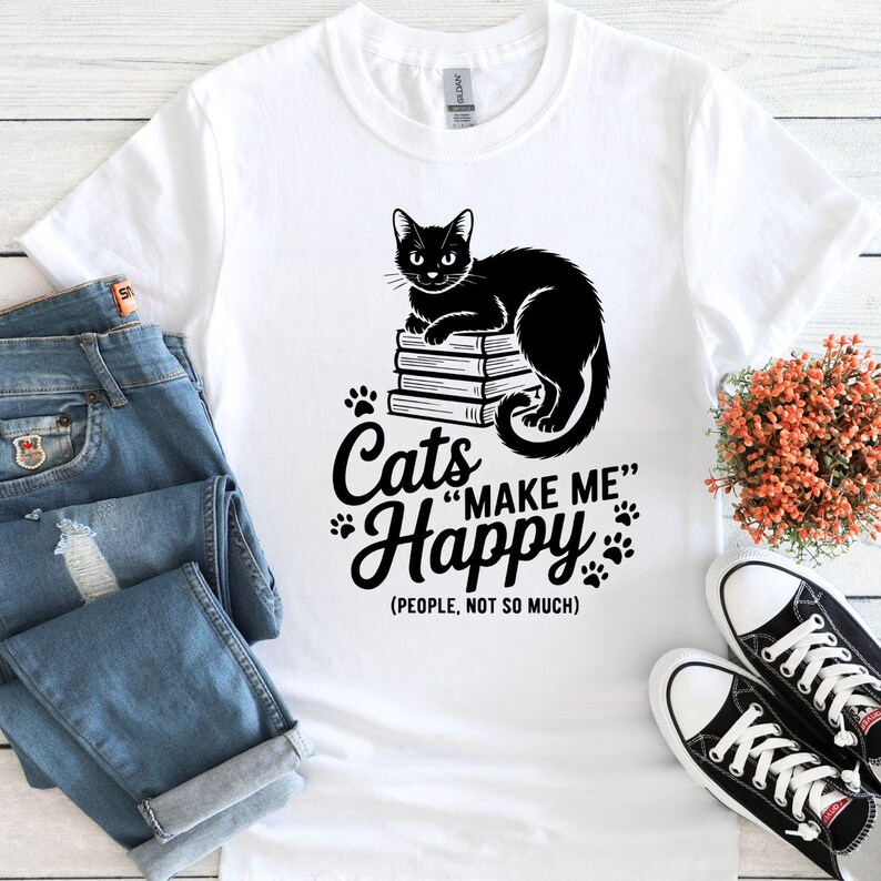 May include: White t-shirt with a black graphic of a cat sitting on books, and the text "Cats Make Me Happy (People, Not So Much)". The shirt is paired with blue jeans and black and white sneakers.