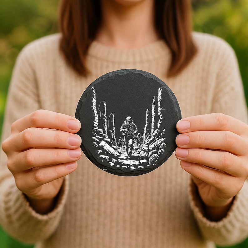 May include: A round, black slate coaster featuring a detailed white illustration of a soldier walking through a war-torn landscape. The design includes architectural ruins and a figure carrying a rifle. The coaster is held up.