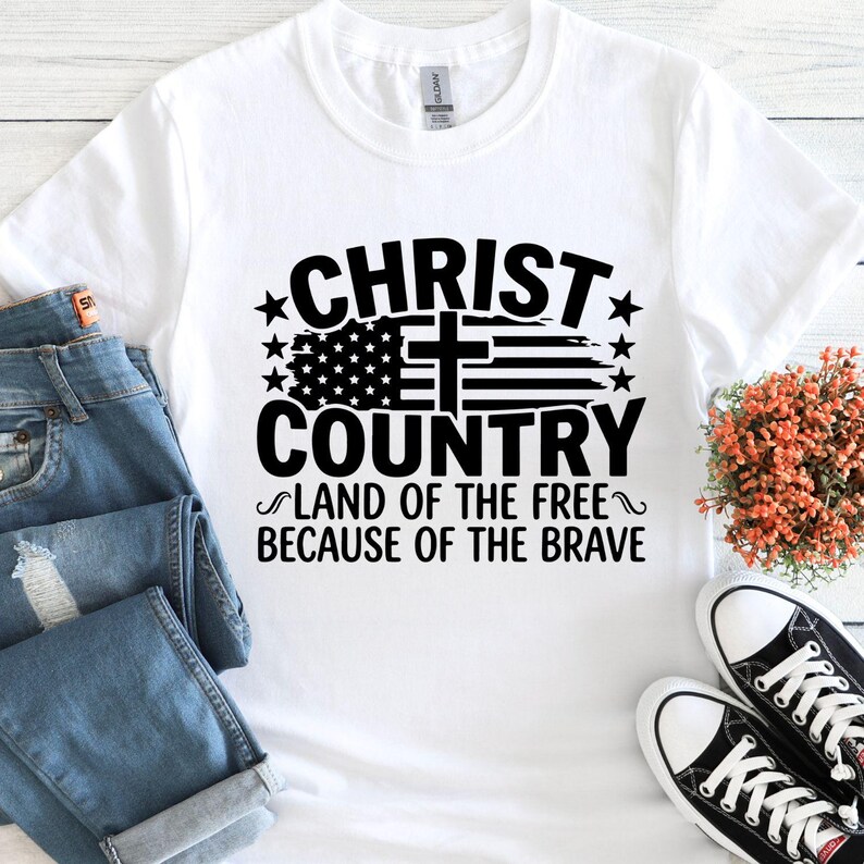 May include: White t-shirt with the text "CHRIST COUNTRY LAND OF THE FREE BECAUSE OF THE BRAVE" in black. The design includes a cross, stars, and a stylized American flag. Blue jeans and black sneakers are also visible.
